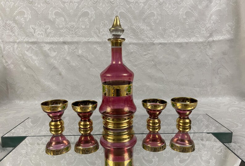 May include: A vintage decanter set featuring a ruby red glass decanter with a gold stopper and accents, accompanied by four matching goblets. The set is displayed on a reflective surface.
