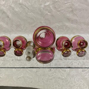 May include: A collection of vintage perfume bottles and a stopper. The bottles are pink with gold rims and accents. The stopper is clear glass. The bottles are arranged on a reflective surface.