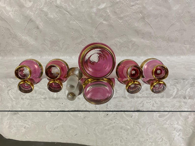 May include: A collection of vintage perfume bottles and a stopper. The bottles are pink with gold rims and accents. The stopper is clear glass. The bottles are arranged on a reflective surface.
