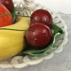 Może przedstawiać: Biała ceramiczna miska na owoce z ozdobnym wzorem ażurowym. Miska zawiera ż&oacute;łty banan, czerwone jabłko i trzy czerwone wiśnie.
