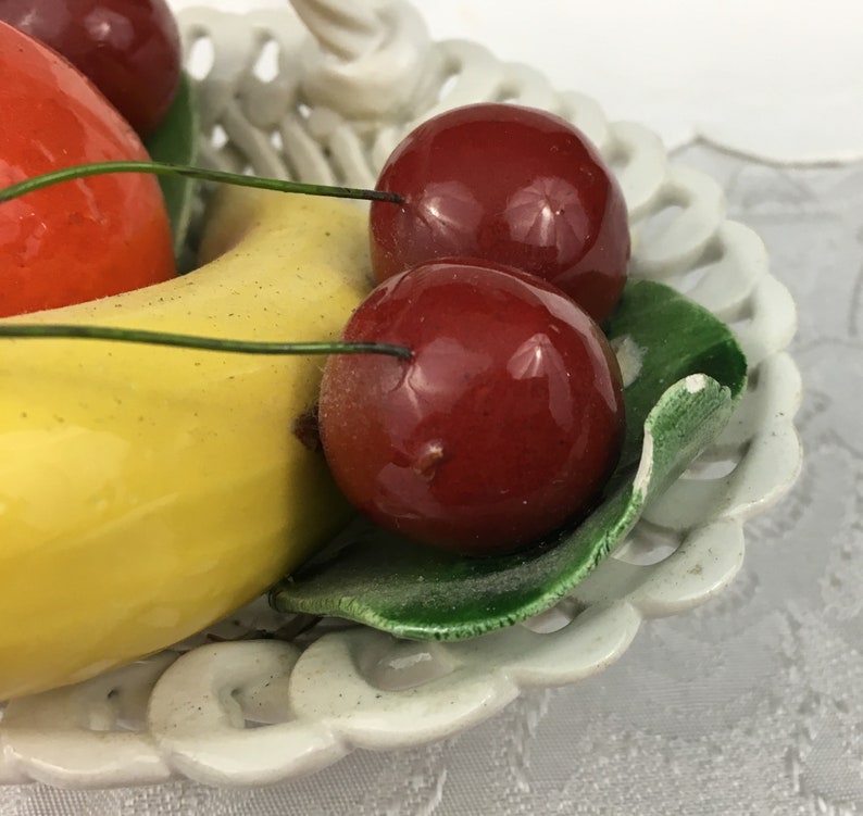 Może przedstawiać: Biała ceramiczna miska na owoce z ozdobnym wzorem ażurowym. Miska zawiera ż&oacute;łty banan, czerwone jabłko i trzy czerwone wiśnie.