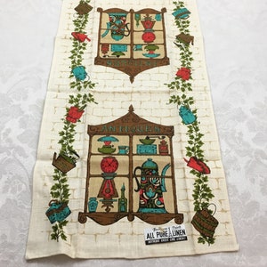 May include: Vintage kitchen tea towel with a white background and a brick pattern. The towel features two storefront windows with the words "Antiques" and "Sewing" above them. The windows are filled with various antique items, including a clock, a teapot, and a coffee grinder. The towel is bordered with green ivy and red flowers.