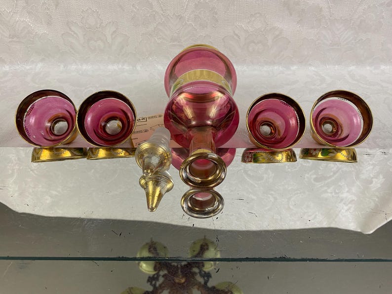 May include: A vintage glass perfume bottle set with a pink and gold design. The set includes a bottle, a stopper, and four matching atomizer tops with gold bases. The bottle and atomizer tops have a swirl pattern.