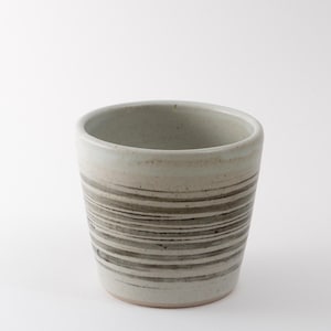 Puede incluir: Un vaso de cerámica con un diseño a rayas grises claras y marrones. La taza tiene forma cilíndrica y un acabado liso sin esmaltar. Las rayas son horizontales y de ancho variable, creando una apariencia texturizada. Adecuado para bebidas.