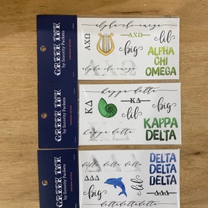 May include: Three sheets of temporary tattoos featuring Greek letters and symbols. The first sheet features a harp and the letters AXO, the second sheet features a seashell and the letters KΔ, and the third sheet features a dolphin and the letters ΔΔΔ.