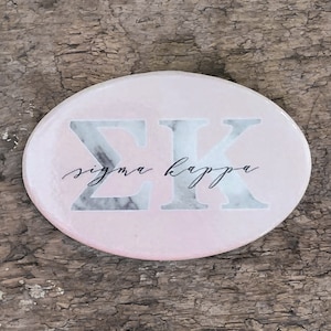 May include: A pink and white marble-patterned round coaster with the letters "ΣΚ" and the text "sigma kappa" in black cursive script.