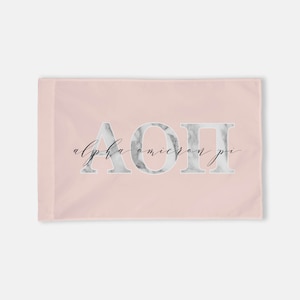 May include: A rectangular, light pink flag featuring the Greek letters ΑΟΠ in a marble-like design. The words "alpha omicron pi" are written in a cursive font below the letters. The flag is a soft pink color.