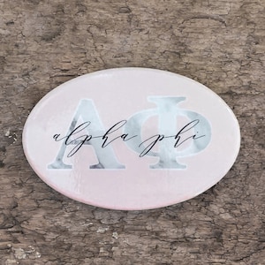 May include: A pink and white marble-patterned oval coaster with the letters "AΦ" and the text "alpha phi" in black script.