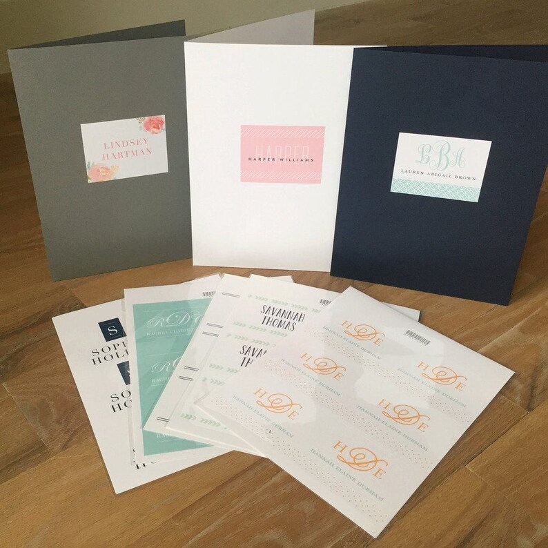 May include: A flat lay of various wedding stationery items, including envelopes, cards, and stickers. The envelopes are gray, white, and navy blue. The cards have different names printed on them, including Lindsey Hartman, Harper Williams, and Lauren Aridail Brown. The stickers have the initials "HDE" printed on them in orange.