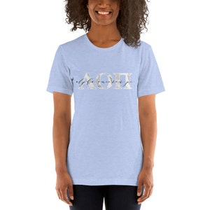 Peut inclure: T-shirt bleu clair avec un motif marbré argenté des lettres grecques "AOPI" et le texte "alpha omicron pi" en écriture noire.