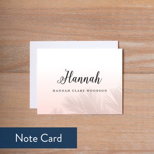 May include: A white note card with a pink and white ombre background featuring a palm leaf design. The name "Hannah" is written in cursive at the top of the card, with the text "Hannah Clare Woodson" below it.