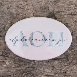 May include: A pink oval-shaped coaster with a marble design and the letters "AOII" in black with a white marble design. The text "alpha omicron pi" is written in black cursive below the letters.