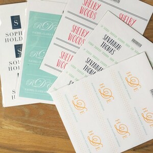 May include: A set of white cards with various names printed on them. The names include Shelby Woods, Savannah Thomas, Rachel Claire, and Hannah Elaine Durham. The cards are decorated with different patterns, including green leaves, gold glitter, and a light blue background.
