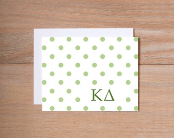 May include: White card with a light green polka dot pattern and the letters KΔ in dark green. The card is placed on top of a white envelope. The background is a light brown wooden surface.