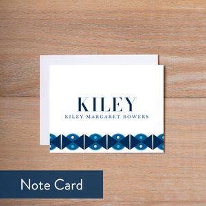 May include: A white note card with a blue and gold geometric border. The text "KILEY" is printed in navy blue at the top, and "KILEY MARGARET BOWERS" is printed in navy blue below the name. The card is on a wood background.