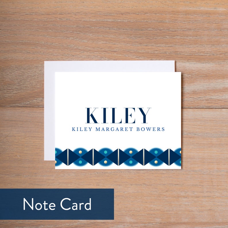 May include: A white note card with a blue and gold geometric border. The text "KILEY" is printed in navy blue at the top, and "KILEY MARGARET BOWERS" is printed in navy blue below the name. The card is on a wood background.