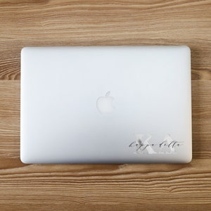 May include: A silver laptop with the Apple logo on the lid. A decal with the Greek letters Kappa Delta and cursive text is on the lower right corner. The laptop is on a wooden surface.