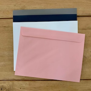 May include: A stack of three envelopes in different colors: gray, navy blue, and pink. The envelopes are all the same size and are lying flat on a wooden surface.