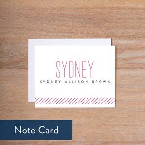 May include: White note card with a pink border and the text "SYDNEY" in a pink sans-serif font. Below the name is the text "SYDNEY ALLISON BROWN" in a smaller pink sans-serif font.
