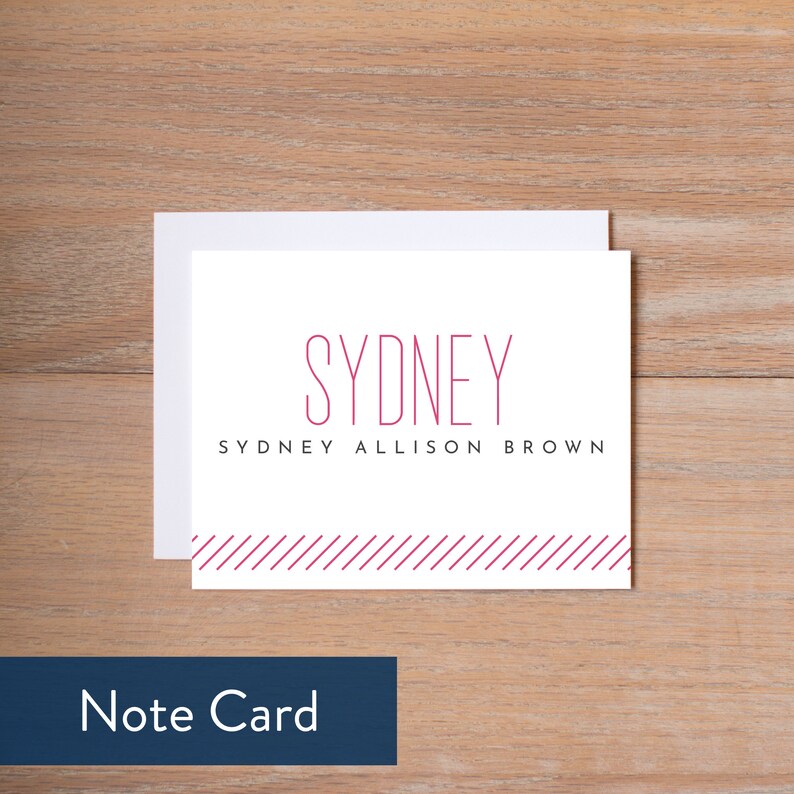 May include: White note card with a pink border and the text "SYDNEY" in a pink sans-serif font. Below the name is the text "SYDNEY ALLISON BROWN" in a smaller pink sans-serif font.