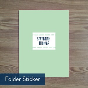 May include: A pale green folder with a white label bearing the name "Savannah Thomas" in navy blue. The label is decorated with light green arrow patterns. The words "Folder Sticker" are in a navy blue box at the bottom.