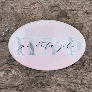 May include: A pink oval coaster with a marble design and the letters "ΠΒΦ" with the words "pi beta phi" written in black cursive.