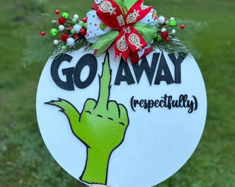 May include: A white round wooden door hanger with a green Grinch hand giving the middle finger and the text "GO AWAY (respectfully)". The door hanger is decorated with a red and green Christmas bow.
