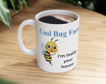 Puede incluir: Taza de cerámica blanca con asa negra. La taza tiene una abeja de dibujos animados con el texto "Cool Bug Facts" y "I'm inside your house." impreso en ella.