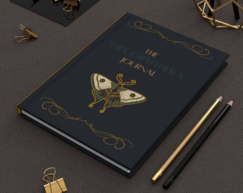 May include: A black journal with gold accents and a gold butterfly design. The text on the cover reads "The Opodiphthera Journal".