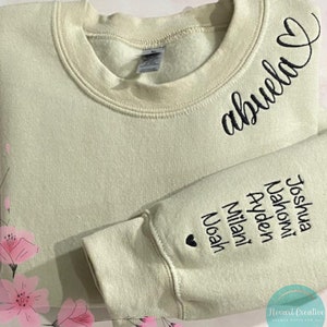 May include: A light green sweatshirt with the word "abuela" and a heart embroidered in black on the front. The sleeve features embroidered names and a heart. A cozy, personalized gift.