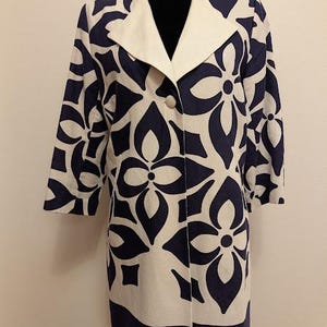 May include: A long, cream-colored coat with a navy blue floral pattern. The coat has a white collar and two buttons. The sleeves are three-quarter length. The coat is displayed on a black mannequin.