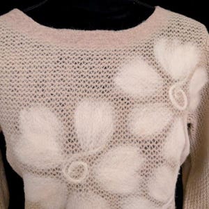 May include: A beige sweater with a floral design. The sweater has a crew neck and long sleeves. The floral design is made of a fuzzy, off-white material and is placed on the front of the sweater.