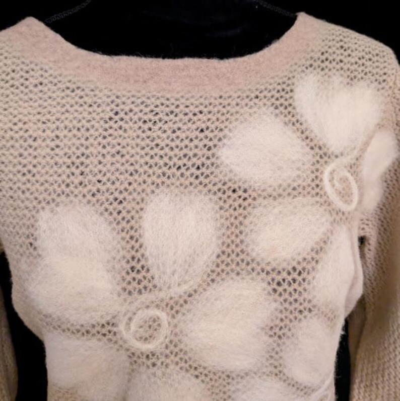 May include: A beige sweater with a floral design. The sweater has a crew neck and long sleeves. The floral design is made of a fuzzy, off-white material and is placed on the front of the sweater.