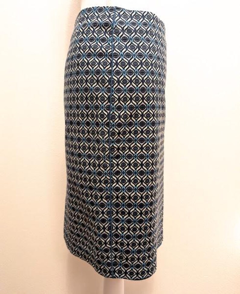 K&ouml;nnte beinhalten: Ein knielanger Rock mit einem geometrischen Muster in Marineblau, Wei&szlig; und T&uuml;rkis. Der Stoff hat einen dezenten Glanz, und das Design weist ein sich wiederholendes Diamanten- und geschwungenes Linienmotiv auf. Der Rock ist figurbetont und hat offenbar einen R&uuml;ckenschlitz.