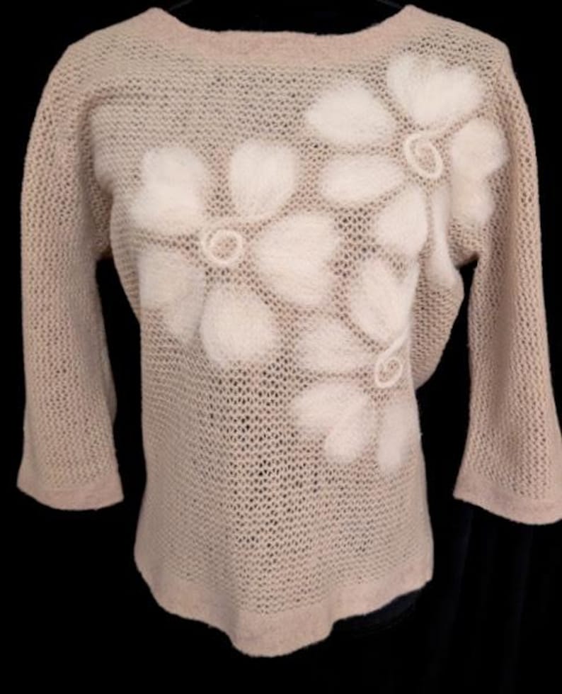 May include: A light beige, open-weave sweater with three-quarter sleeves. The sweater features large, white, fluffy flower appliqu&eacute;s across the front. The neckline, cuffs, and hem are a solid beige.