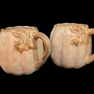 May include: Two ceramic mugs shaped like pumpkins with a glossy cream-colored finish. Each mug features a sculpted brown handle resembling a vine and a decorative leaf detail near the top rim, with vertical ridges mimicking a pumpkin's texture.