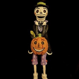 May include: A standing skeleton figurine wearing a black top hat with a green band, a black glittery coat, and purple pants with a diamond pattern. The skeleton holds a round orange pumpkin with a carved face and wears white shoes.