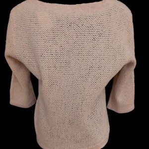 May include: A light pink, open-knit sweater with a boat neck and three-quarter sleeves. The sweater has a relaxed fit and a slightly textured appearance. The sleeves are wide and taper towards the elbow.
