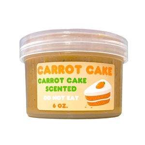 May include: A clear plastic jar filled with a brown, creamy substance. The jar has a label that reads "Carrot Cake Carrot Cake Scented Do Not Eat 6 oz.". The label also features a cartoon image of a slice of carrot cake with a carrot on top.
