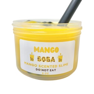 May include: A clear plastic container filled with yellow slime with black boba balls. The container has a white label that reads "Mango Boba Mango Scented Slime Do Not Eat 6 oz."