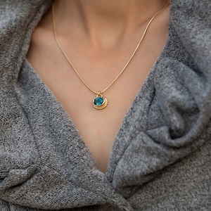 Może przedstawiać: Złoty łańcuszek z niebieskim kamieniem szlachetnym wisiorkiem. Wisiorek jest osadzony w złotej ramie z fakturowanym brzegiem.