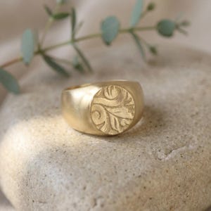 A gold ring with a carved leaf design sits on a rock, with a branch of greenery in the background.