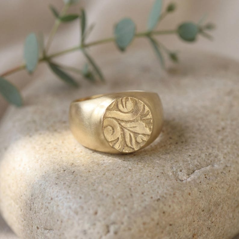 A gold ring with a carved leaf design sits on a rock, with a branch of greenery in the background.