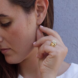 A close-up of a woman's face, with her hand gently touching her ear and a gold ring visible on her finger.