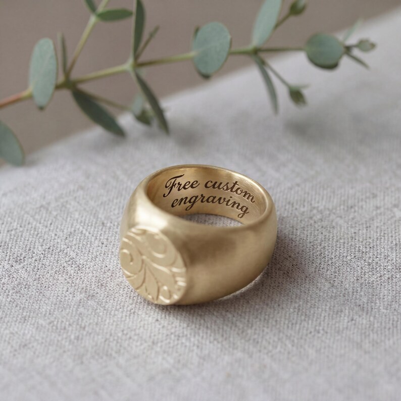 A gold ring with a carved design sits on a textured surface, accompanied by a branch of green leaves.