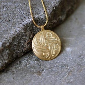 May include: A gold-colored pendant necklace with a circular pendant featuring an embossed spiral design. The pendant hangs from a thin, gold chain. The necklace is displayed on a gray, textured surface.