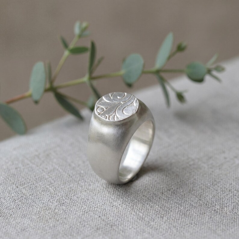 A silver ring with a floral design sits on a textured gray surface, with a branch of green leaves in the background.
