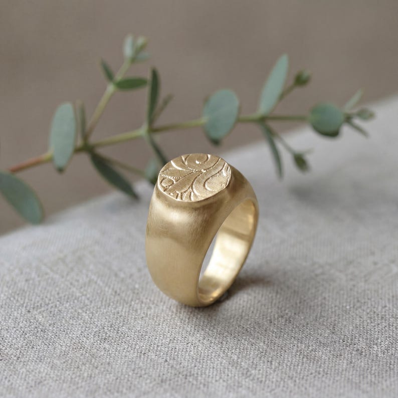 Gold floral signet ring with a wide band and engraved botanical pattern, displayed on soft linen with a blurred green branch in the background.