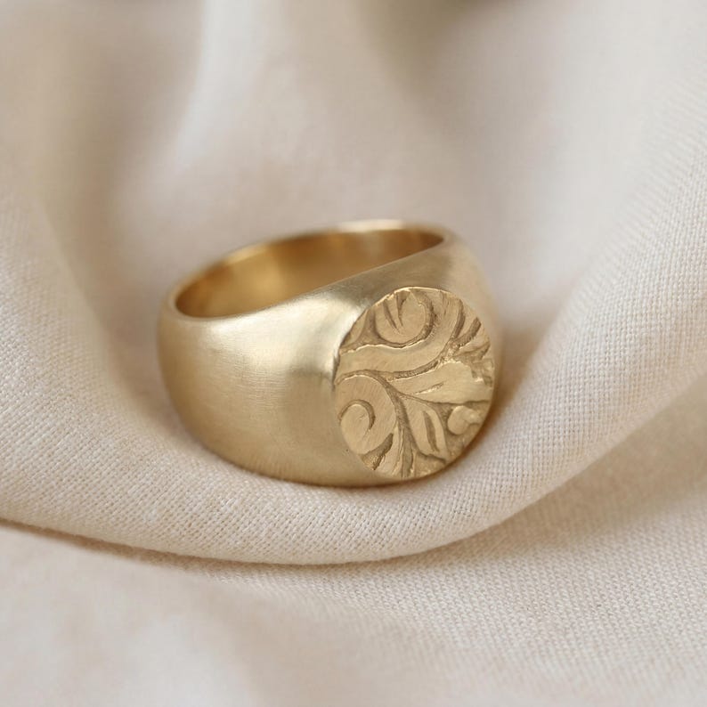 A gold ring with a circular embossed design is resting on a white fabric surface.