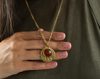 Puede incluir: Un collar de oro con un colgante redondo que presenta una piedra preciosa roja y detalles en forma de hoja.